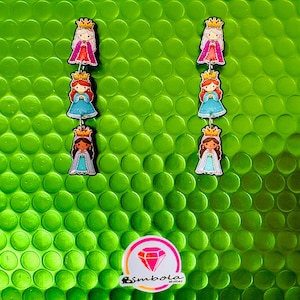 May include: Three princess-themed charm earrings. Each charm features a princess with a crown, wearing a dress in shades of pink, blue, and white. The earrings are displayed against a textured green background, with a logo in the bottom right corner.