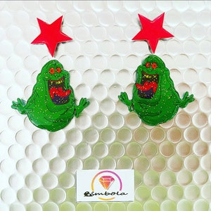 May include: A pair of green glitter ghost earrings with red star charms. The earrings are on a white background with a bubble wrap texture.