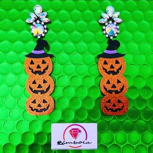 Puede incluir: Un par de pendientes con temática de Halloween. Cada pendiente presenta una pila de tres calabazas naranjas brillantes con caras sonrientes, rematadas con un sombrero de bruja negro con una banda morada. Los pendientes están adornados con cristales iridiscentes.