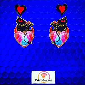 May include: Two black devil figures with red eyes and tails are sitting on pink and blue hearts with flames. The hearts are outlined in gold and have a gold chain. The figures are holding red hearts above their heads. The background is a bright blue.
