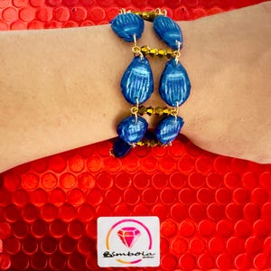 May include: A blue bracelet with teardrop-shaped beads and gold-colored accents. The bracelet is made of multiple blue beads connected by gold-colored chains. The bracelet is on a wrist, with a red background. The logo of the brand "Bimbola" is visible.