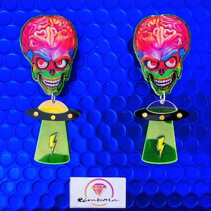 Puede incluir: Un par de pendientes con una cabeza de extraterrestre verde con un cerebro rosa y un platillo volante amarillo con un rayo. Los pendientes están sobre un fondo azul.