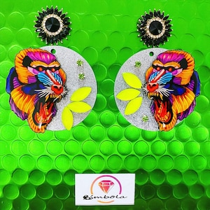 Puede incluir: Un par de pendientes de plata con un diseño de mandril de mono colorido y un adorno de pedrería negra. Los pendientes están sobre un fondo verde.