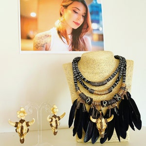 Puede incluir: Un collar negro y dorado con plumas negras y un colgante de calavera. El collar está formado por varias capas de cuentas negras y doradas y tiene un estilo tribal.