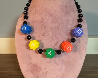 May include: A necklace with black beads and colorful, flower-shaped pendants. The pendants are blue, yellow, green, and red, each with a different white symbol: circle, triangle, diamond, and circle.