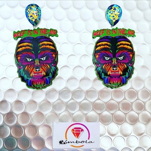 Puede incluir: Un par de pendientes coloridos con un diseño de cara de hombre lobo y el texto "Wolf is in the air". Los pendientes están sobre un fondo blanco con textura de plástico de burbujas.