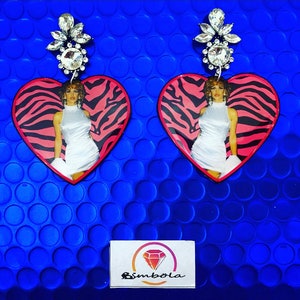 Puede incluir: Un par de pendientes en forma de corazón con un diseño de cebra rosa y negro. Cada pendiente presenta una foto de una mujer con un vestido blanco y un cúmulo de diamantes de imitación en la parte superior.