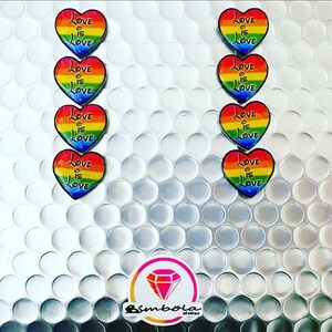 Puede incluir: Diez pegatinas en forma de corazón arcoíris con el texto "Love is Love" en cada una. Las pegatinas están dispuestas en dos columnas de cinco.