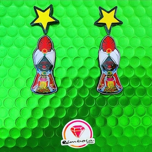 May include: A pair of earrings featuring a cartoon character wearing a red and white turban and a gold and red robe. The earrings are topped with a yellow star.