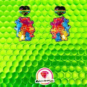 Op de afbeelding: Een verzameling kleurrijke stickers en pins op een heldergroene achtergrond. De stickers tonen cartoonfiguren in verschillende kleuren, en de pins zijn hartvormig met het woord "Fuzzies". Een logo met een diamant is ook zichtbaar.