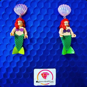 May include: A pair of earrings featuring a mermaid Lego figurine with a green tail and a seashell charm. The mermaid is wearing a pink bikini top. The earrings are on a blue background with a small logo that says "Simbola" in a diamond shape.