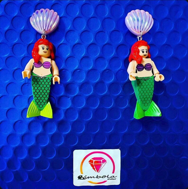 May include: A pair of earrings featuring a mermaid Lego figurine with a green tail and a seashell charm. The mermaid is wearing a pink bikini top. The earrings are on a blue background with a small logo that says "Simbola" in a diamond shape.