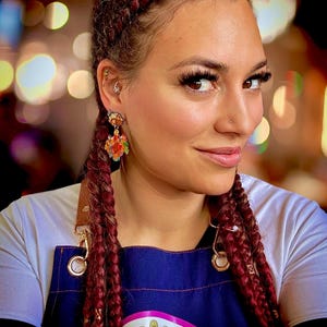 May include: A woman wearing dangling earrings with a colorful design. The earrings feature a cartoon-like illustration. The woman has long, braided red hair and is wearing a blue apron with a cartoon graphic.