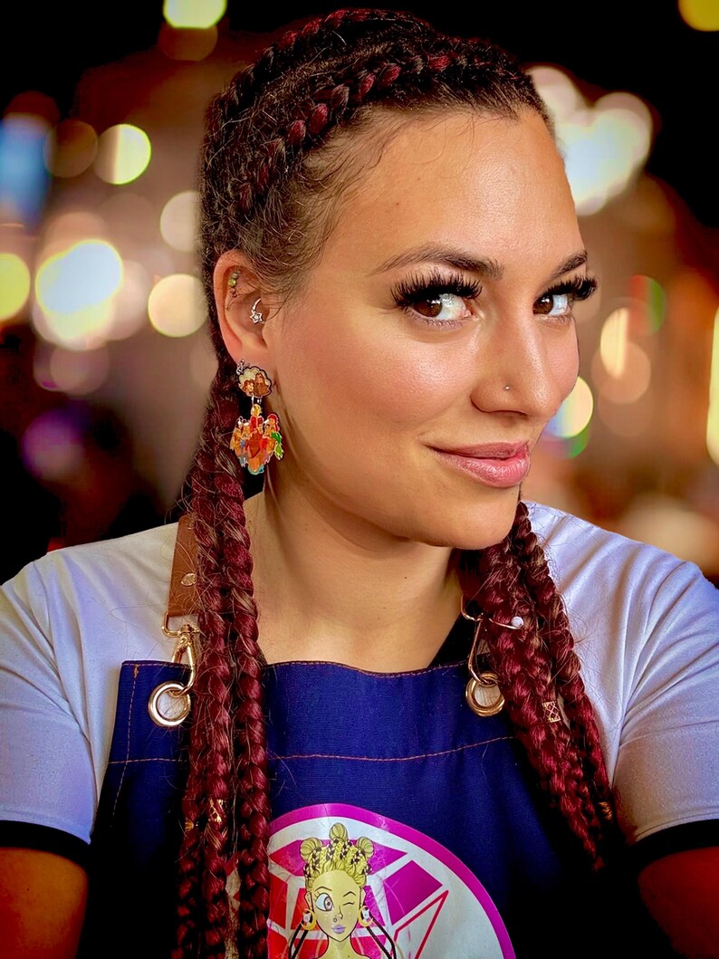 May include: A woman wearing dangling earrings with a colorful design. The earrings feature a cartoon-like illustration. The woman has long, braided red hair and is wearing a blue apron with a cartoon graphic.