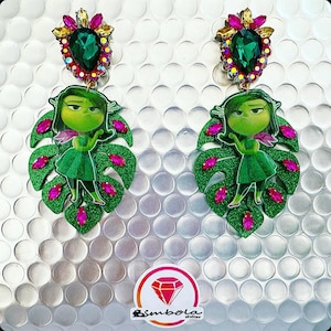 Puede incluir: Un par de pendientes verdes y rosas con un personaje de dibujos animados con la cara verde y un vestido verde. Los pendientes tienen forma de hoja verde con detalles rosas. Los pendientes están decorados con diamantes de imitación.