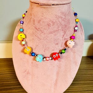May include: A colorful beaded necklace featuring Pac-Man and ghost character beads. The necklace includes a variety of colorful beads in shades of yellow, blue, red, pink, and green. The necklace is displayed on a pink jewelry bust.