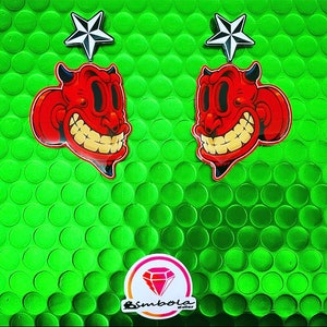 May include: Two red devil head earrings with a white star charm on each. The earrings are on a green background.