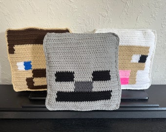 May include: Three square crochet pillows featuring pixelated character faces. The left pillow is tan and brown with blue eyes. The center pillow is gray with black eyes and mouth. The right pillow is white and tan with a pink nose.