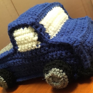 May include: A handmade crochet toy truck featuring a deep blue body, white windows, black tires with gray centers, and a gray truck bed. The yarn construction shows a textured stitch pattern throughout the entire vehicle shape.