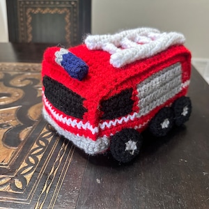 May include: A red and gray crocheted fire truck toy with a white ladder on top. The truck has black wheels and a blue light on the front.