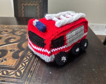 May include: A red and gray crocheted fire truck toy with a white ladder on top. The truck has black wheels and a blue light on the front.