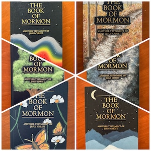 May include: Four different covers of the Book of Mormon, each with a unique painted design. The covers feature a rainbow, a tree, butterflies and flowers, a path through a forest, and a moonlit mountain range.