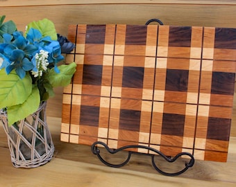 May include: A rectangular wooden cutting board with a plaid pattern in shades of brown and tan, displayed on a black metal stand. A small bouquet of blue and white flowers in a wicker basket is to the left.