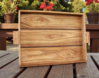 May include: A rectangular wooden serving tray with a natural finish. The tray has a light brown color with darker brown accent lines and handles on each side. The tray is sitting on a wooden table.