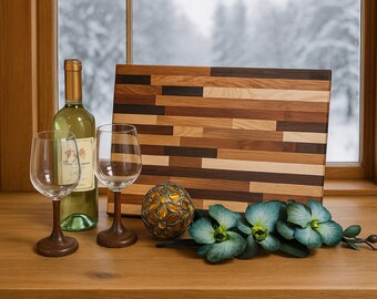 May include: A still life arrangement featuring a bottle of white wine, two wine glasses, a wooden cutting board, a decorative orb, and blue-green orchids. The cutting board is made of various wood tones. The scene is set on a wooden surface near a window.