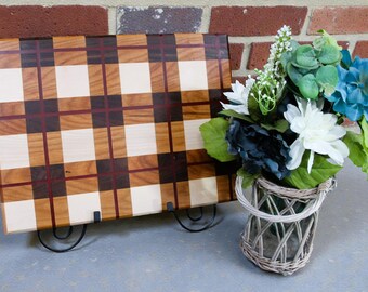 May include: A wooden cutting board with a plaid pattern in shades of brown, tan, and red. The board is propped up on a black metal stand. A small woven basket holds a bouquet of blue, white, and green flowers.
