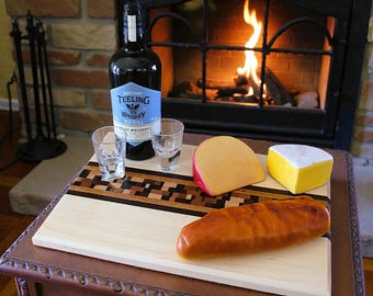 May include: A wooden serving board with a decorative inlaid design holds a bottle of Teeling Whiskey, two shot glasses, and a selection of cheeses and a baguette. The scene is set in front of a fireplace with a burning fire.