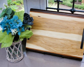 May include: A rectangular wooden serving tray with dark brown handles and trim. The tray has a light wood grain pattern with a darker wood border. A vase of blue and green flowers sits to the left of the tray.