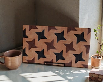 May include: A rectangular wooden cutting board with a star pattern in shades of brown and black. A brown ceramic mug sits to the left. A small potted plant is to the right. The cutting board measures approximately 18 inches long.