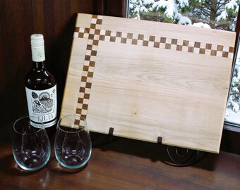 May include: A wooden cutting board with a checkerboard design, a bottle of wine, and two stemless wine glasses. The cutting board is light-colored wood with a dark wood border. The wine bottle has a white label.
