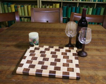May include: A wooden cutting board with a checkerboard pattern, a wine bottle, two wine glasses, and a candle with floral accents are arranged on a wooden table. The wine glasses have dark brown stems.