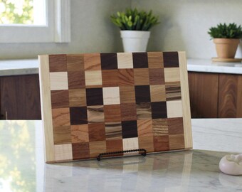 May include: A rectangular wooden cutting board with a checkerboard pattern made of various wood tones, including light, medium, and dark browns. The board is displayed on a white countertop. The board measures approximately 12 inches by 16 inches.