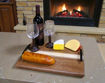 May include: A wooden serving tray with a bottle of red wine, two wine glasses, and two types of cheese. A loaf of bread is also on the tray. The tray has black handles and a light wood stripe.