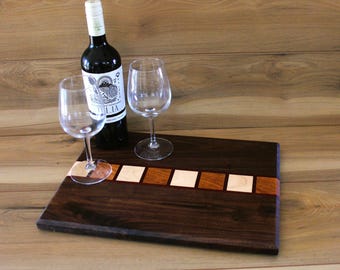May include: A dark wood serving board with a decorative inlaid stripe of light and medium-toned wood squares. A bottle of wine and two wine glasses are arranged on the board, ready for serving.