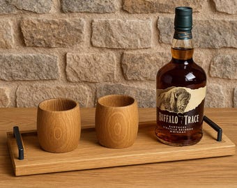 May include: A wooden serving tray with two wooden cups and a bottle of Buffalo Trace Kentucky Straight Bourbon Whiskey. The tray has black handles and is set against a brick wall.