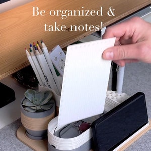 Pode incluir: Um organizador de mesa bege com compartimentos para canetas, lápis, um telefone e uma pequena planta. O organizador tem um design branco e cinza. O texto "Be organized & take notes!" é visível na imagem.