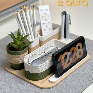 Peut inclure: Un organisateur de bureau blanc et vert avec des compartiments pour stylos, crayons, une télécommande, des lunettes et un téléphone. L'organisateur a une base beige et un support de téléphone blanc et vert avec l'heure 12:28 affichée à l'écran.