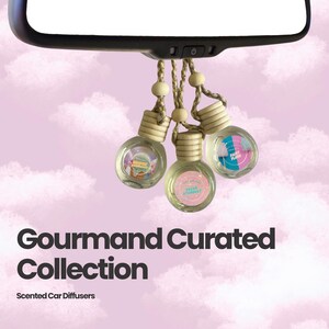 May include: Three small, round glass car diffusers with wooden caps hang from a rearview mirror by braided cords with wooden beads. The labels read Sugar Stardust and Fairy Floss. The text Gourmand Curated Collection Scented Car Diffusers is visible.