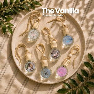 May include: Six round glass car air freshener bottles with wooden caps and braided hanging cords arranged on a circular tray. Each bottle features a unique label with text including The Vanilla Curated Bundle, Vanilla Noir, and other scent names.