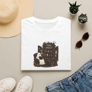 May include: A white t-shirt featuring a graphic of a ghost sitting in an armchair reading a book in front of a dark wooden bookcase. The bookcase has the text "TRICK OR SPEECH" printed on the top arch, surrounded by small pumpkins and candles.