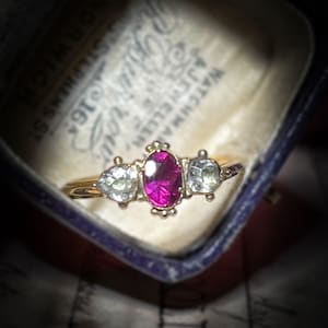 Op de afbeelding: Een gouden ring met een grote ovale robijn-edelsteen omringd door twee kleinere ronde witte edelstenen.