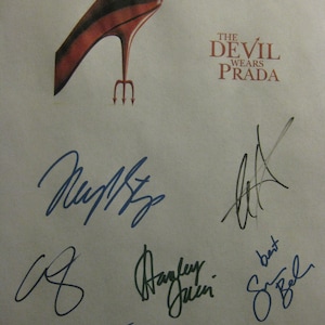 May include: A white sheet of paper with the title "The Devil Wears Prada" and a red high-heeled shoe with a trident for a heel. The paper is signed by several people, including "Meryl Streep" and "Anne Hathaway".