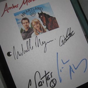 May include: A white page with black and blue signatures from the cast of the TV show Heartland. The signatures include Amber Marshall, Michelle Morgan, Shaun Johnston, and the show's creators. The page also features a photo of the show's logo and a horse.