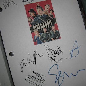 May include: A white sheet of paper with several signatures in black and blue ink. The signatures are from the cast of the movie Jojo Rabbit. The movie poster is in the center of the page. The poster is red and features the title "Jojo Rabbit" in white letters. The cast includes Roman Griffin Davis, Thomasin McKenzie, Scarlett Johansson, Taika Waititi, and Sam Rockwell.
