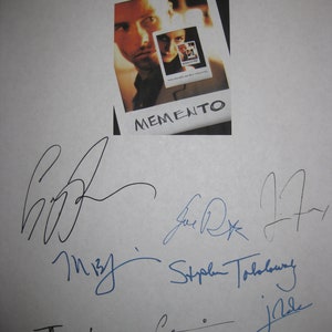 May include: A white sheet with a small movie poster of "Memento" and multiple signatures in black and blue ink. The poster features a man's face and the film's title. The signatures are of various sizes and styles.