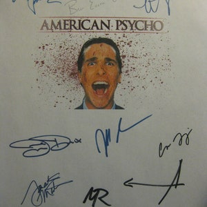 May include: A white poster with the title "American Psycho" in black text. The poster features a close-up of an actor with blood splattered on his face. The poster is signed by several people, including the actor, Willem Dafoe, and the director, Mary Harron.